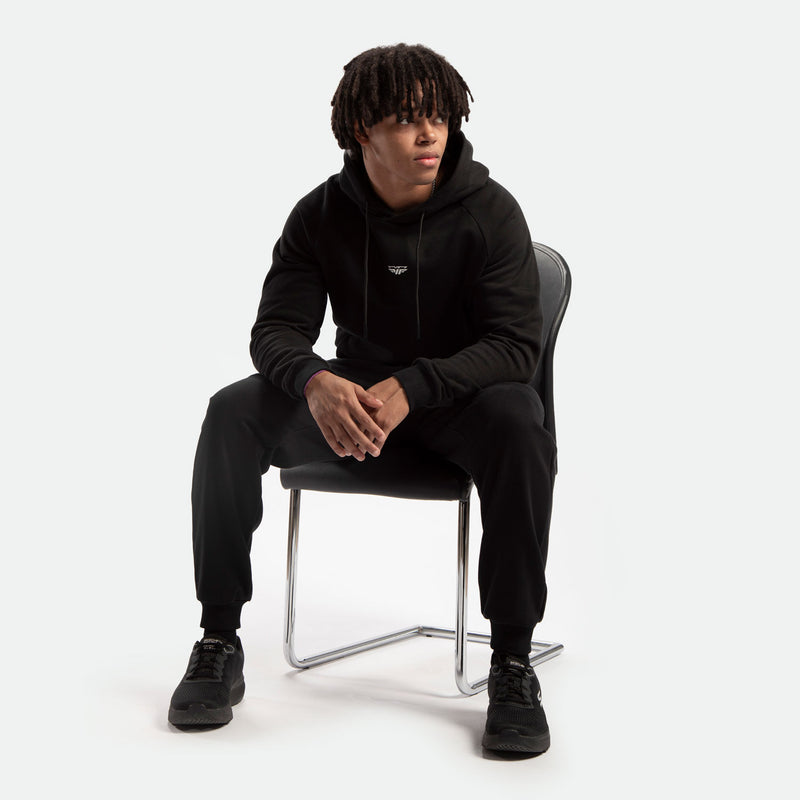 A person with short dreadlocks sits on a chair, wearing a black hoodie and matching pants. They appear relaxed and contemplative, looking to the side.