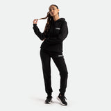 a fit woman wearing matching hoodie and pant with logo ,she wearing  black shoes athletic, plus the background is light grey 