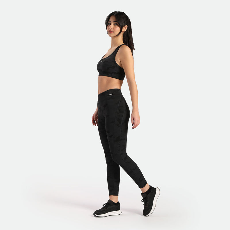 A woman in a black sports bra and leggings stands in profile, showcasing an athletic look against a plain background.