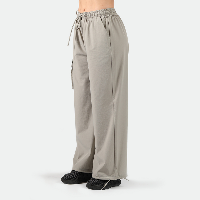 a fit woman stand at right and wearing parachute pants with black shoes athletic ,plus the background is light grey 