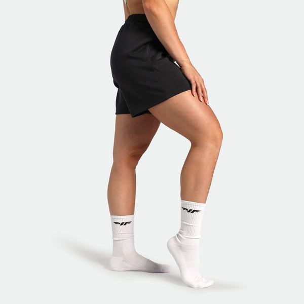 Person wearing black athletic shorts and white socks with a brand logo on a light gray background