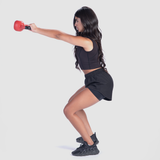 A woman dressed in a black crop top, black athletic shorts, and black sneakers is performing a kettlebell exercise. She is in a squat position, holding a red kettlebell with both hands extended straight in front of her. Her long dark hair is loose