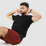Man in black t-shirt and red shorts doing sit-ups against a plain white background. His expression is focused, conveying determination.