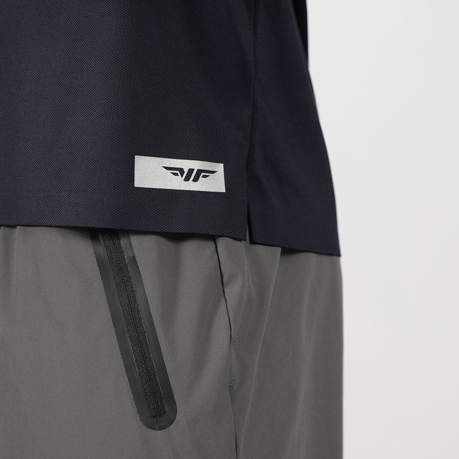 The image shows a close-up of a person wearing a dark blue shirt with a small logo above the hem and gray pants featuring a black zipper on the pocket.