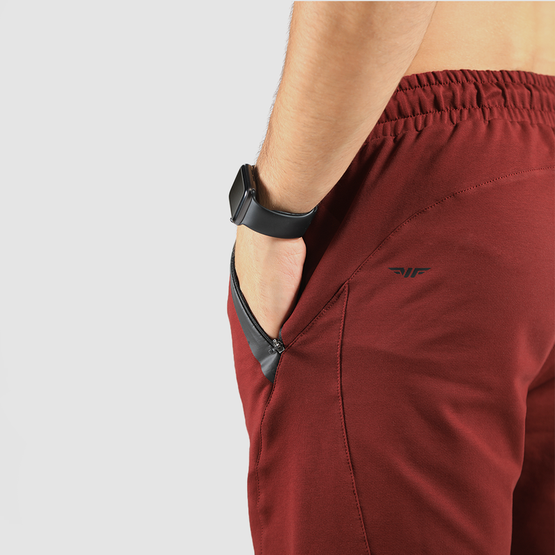 Close-up of a person wearing maroon athletic shorts with a hand in the black zippered pocket. A smart watch is visible on the wrist, against a white background.