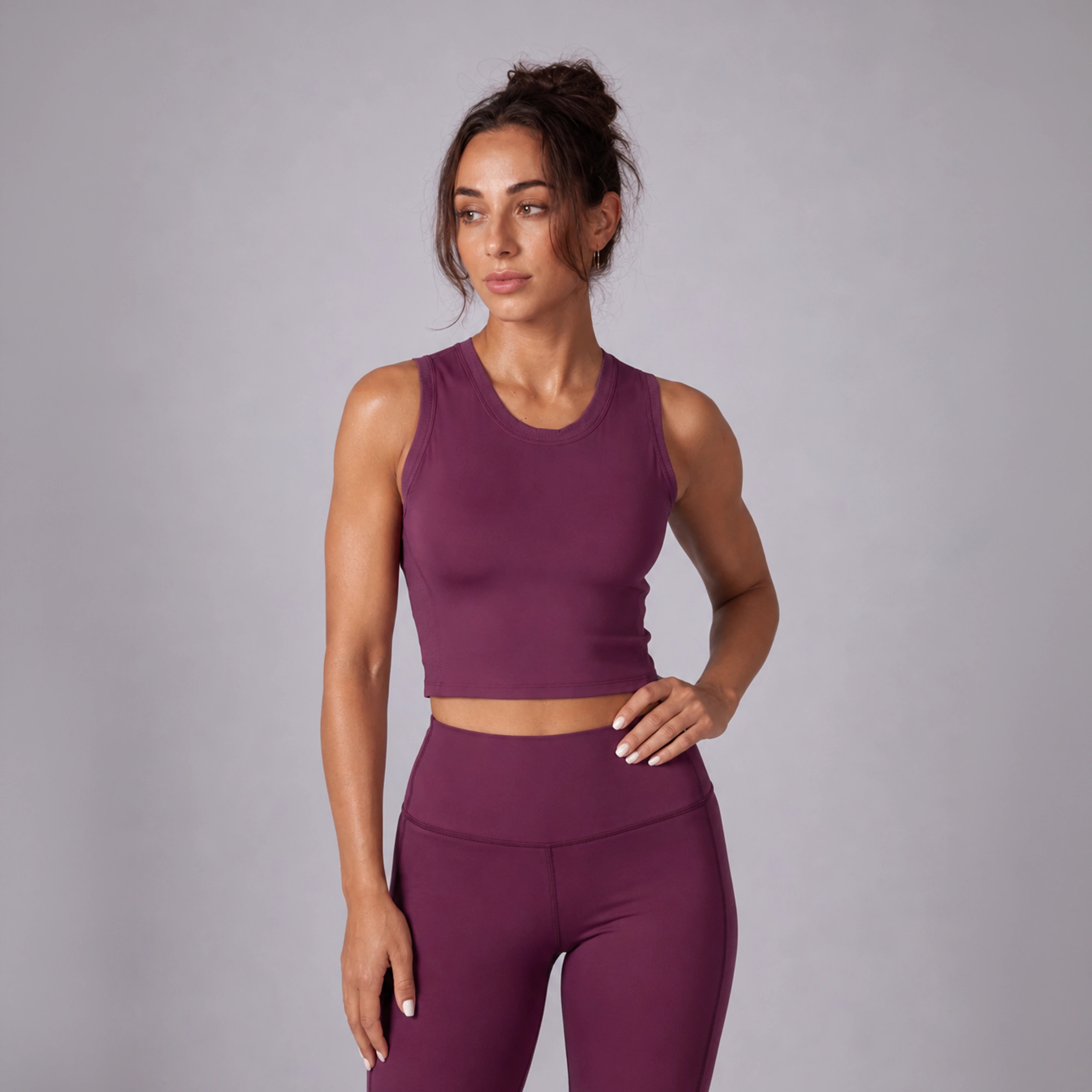 A woman modeling a matching sleeveless crop top and high-waisted leggings set in a deep plum purple color, photographed against a neutral gray studio background.