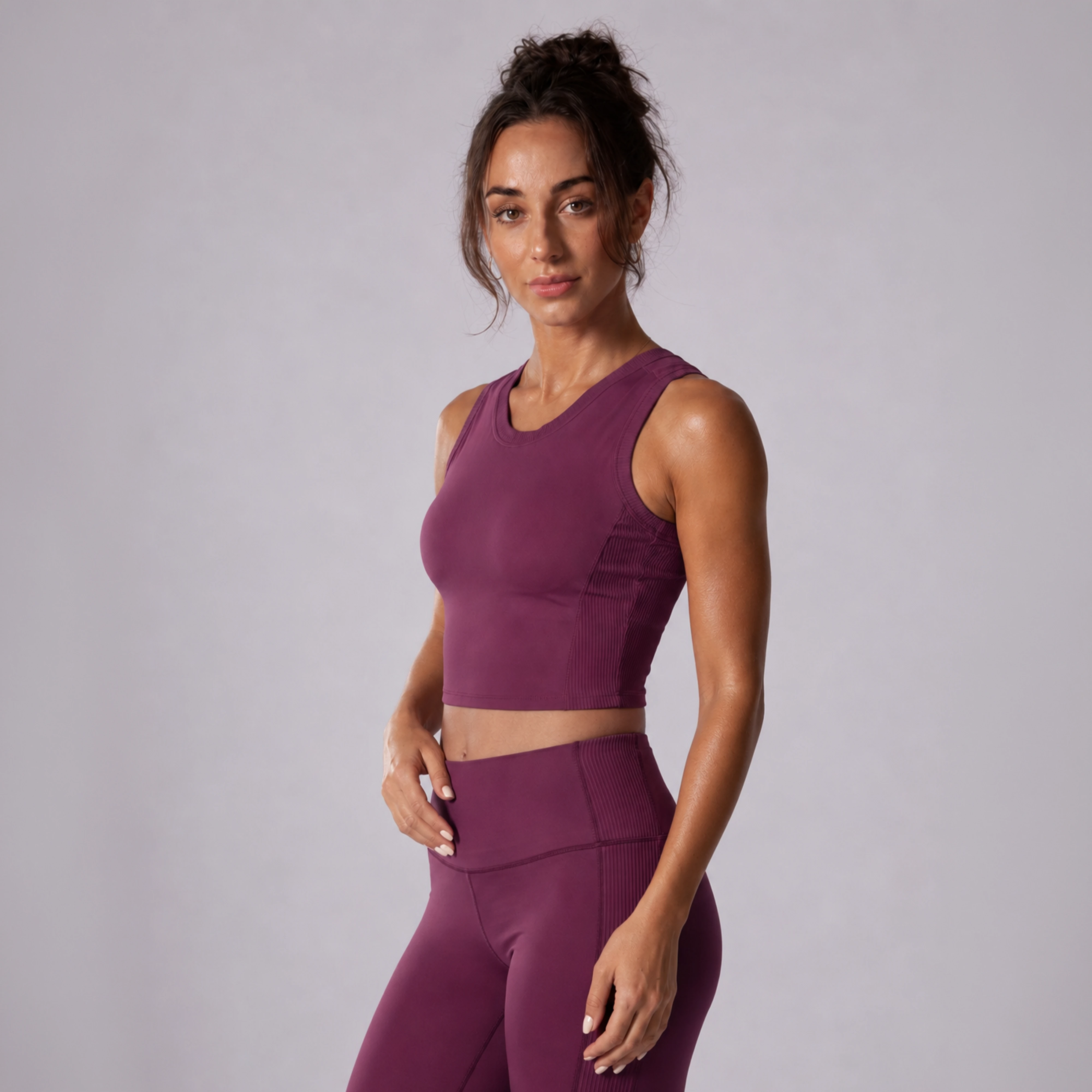 A woman modeling a matching athletic set consisting of a sleeveless scoop-neck crop top and high-waisted leggings in a deep plum purple color, set against a plain light gray studio background.