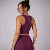 A woman wearing a matching athletic crop top and high-waisted leggings in a deep plum purple, shown from the back against a light gray studio background.