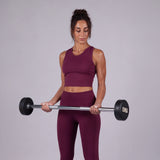 A woman in a matching burgundy activewear set holds a barbell with black weight plates marked '5KG' against a solid light gray studio background.