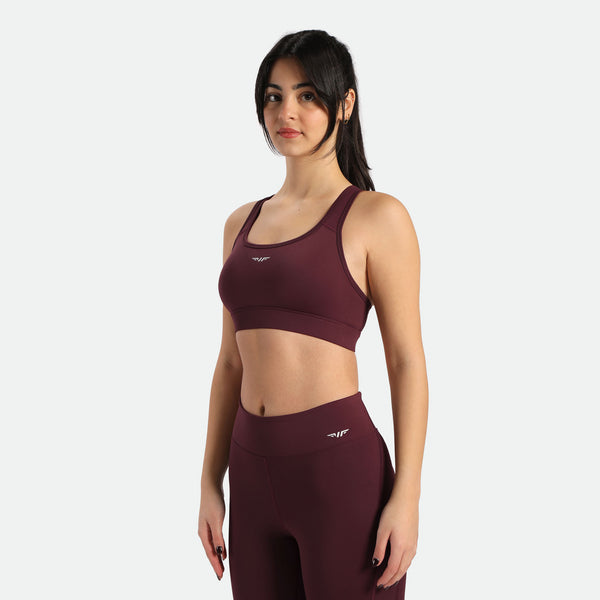 A woman in a dark red sports bra and matching leggings stands in a three-quarter pose, the bra and legging have the same winneforce logo white In different directions, the background is white