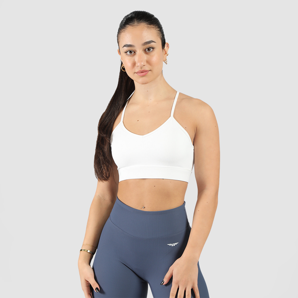 A graceful woman stands confidently in a white sports bra and dark blue high-waisted pants against a plain background