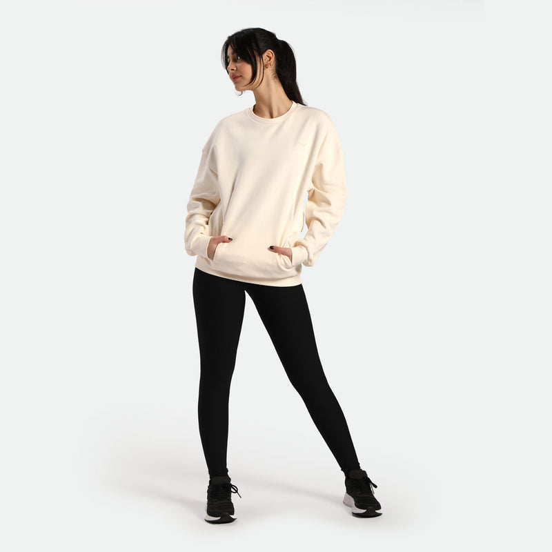 A model wearing a light cream oversized sweatshirt with pockets and black leggings, posing confidently against a white background.