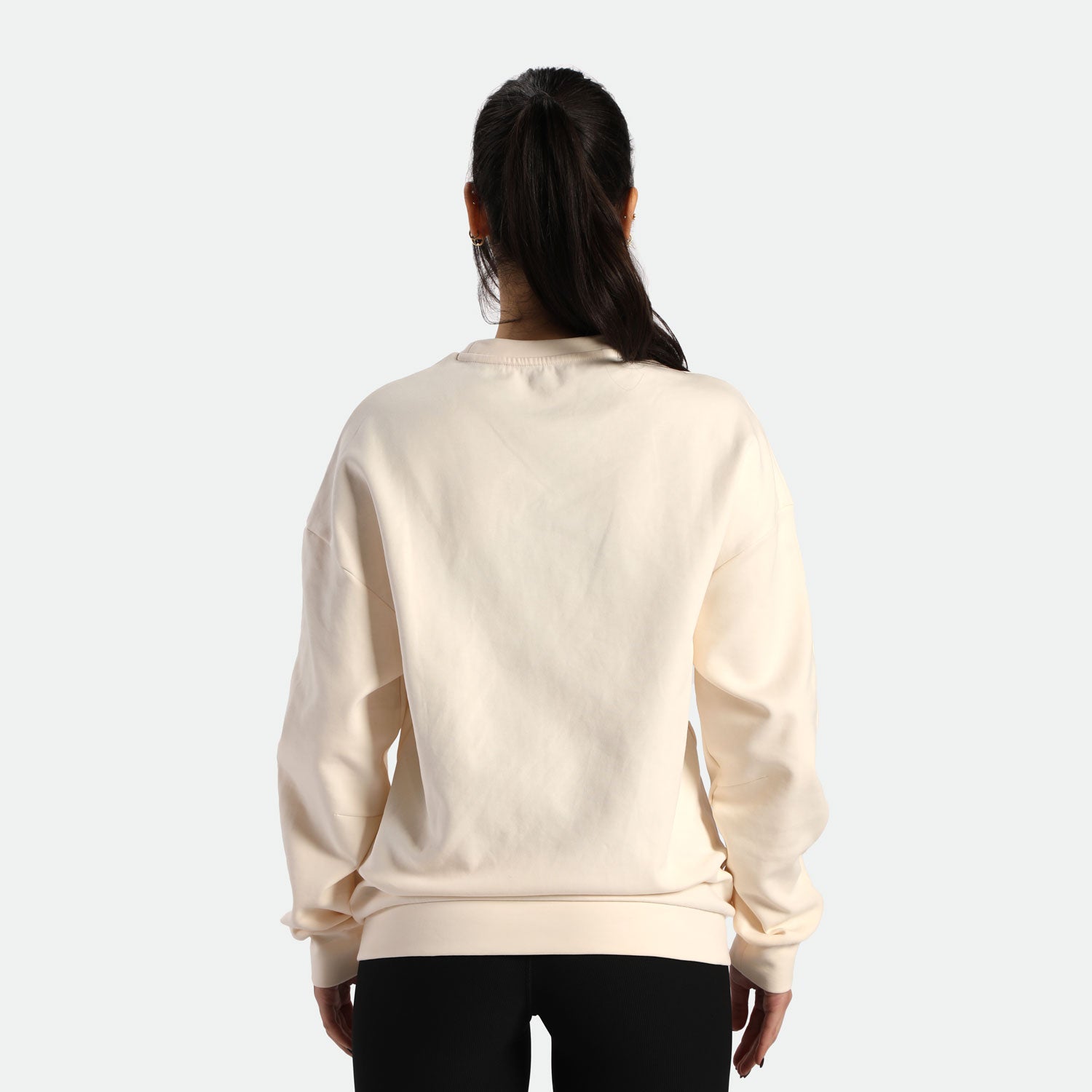 A woman with long hair stands facing away, wearing a light beige sweatshirt and black leggings against a plain background.