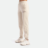 a fit woman stand at right  and she wearing jogger pant beige with white shoes ,plus the background is light grey 