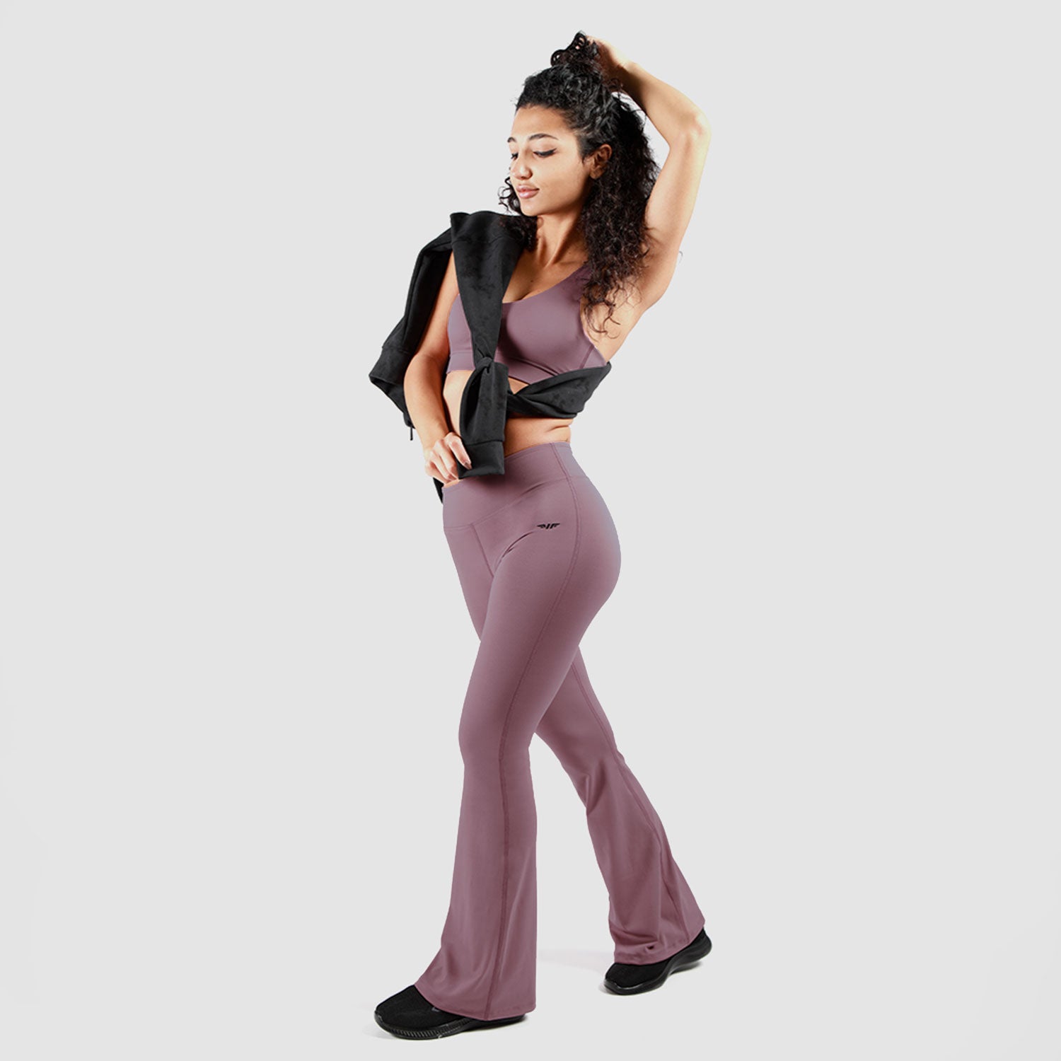 A woman wearing a mauve-colored seamless workout set, including a sports bra and high-waisted flare leggings, posing confidently with a black jacket draped over her shoulder against a neutral gray background. She is also wearing black athletic shoes