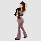 A woman wearing a mauve-colored seamless workout set, including a sports bra and high-waisted flare leggings, posing confidently with a black jacket draped over her shoulder against a neutral gray background. She is also wearing black athletic shoes