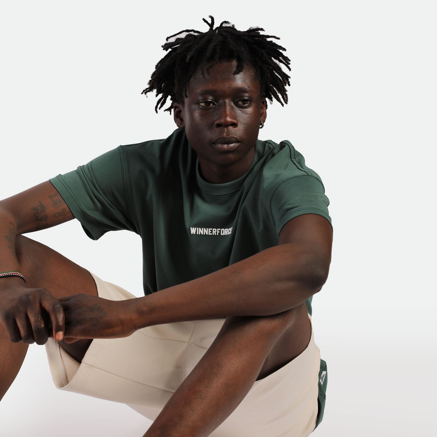 A person sits pensively against a white background, wearing a dark green shirt with "WINNERFORCE" text, and beige shorts. The mood is contemplative.