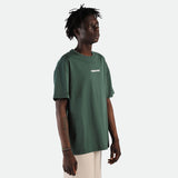 a fit man stand at right  and wearing  t-shirt green  with pants beige 