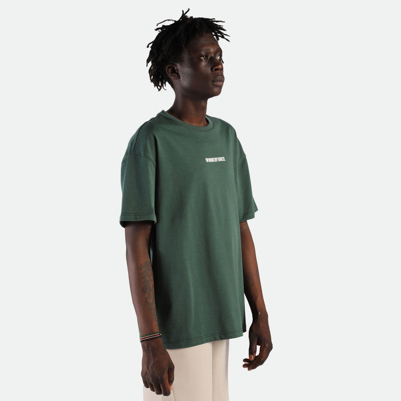 a fit man stand at right  and wearing  t-shirt green  with pants beige 