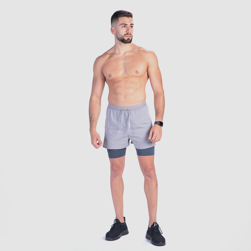 A shirtless man stands confidently against a white background, wearing gray shorts, black sneakers, and a smartwatch. His expression is focused and composed.