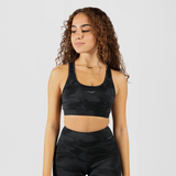 A fit woman with a confident expression poses in a  camo athletic outfit consisting of a sports bra and high-waisted leggings. She stands against a plain white background with her hands resting on her waist.
