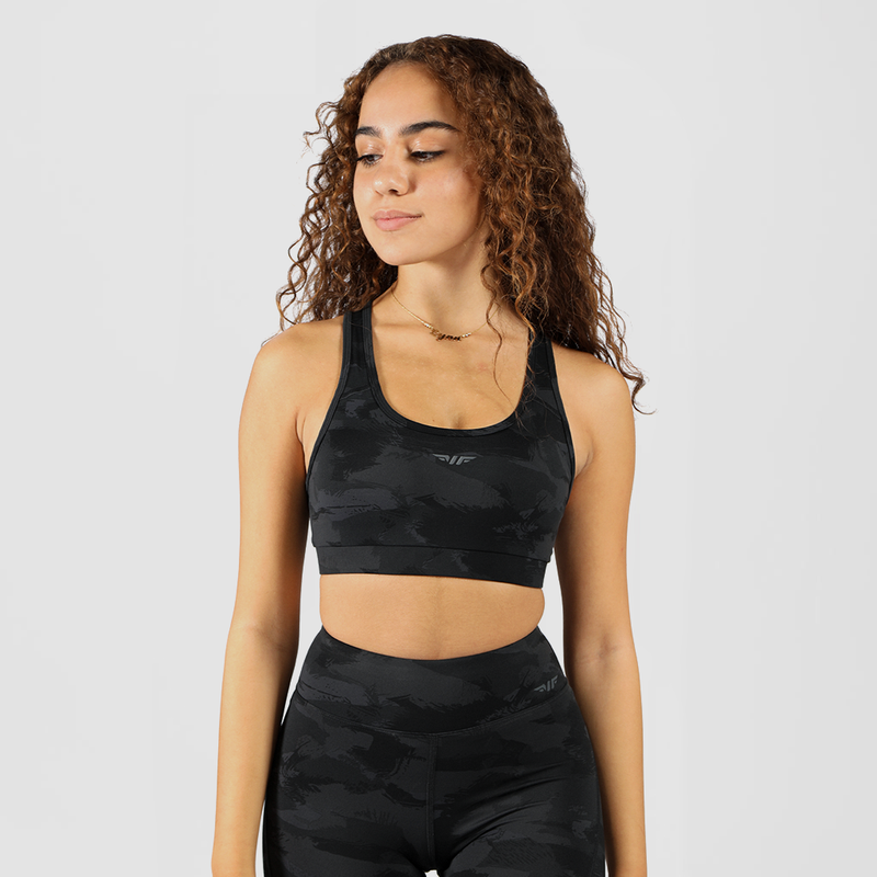 A fit woman with a confident expression poses in a  camo athletic outfit consisting of a sports bra and high-waisted leggings. She stands against a plain white background with her hands resting on her waist.