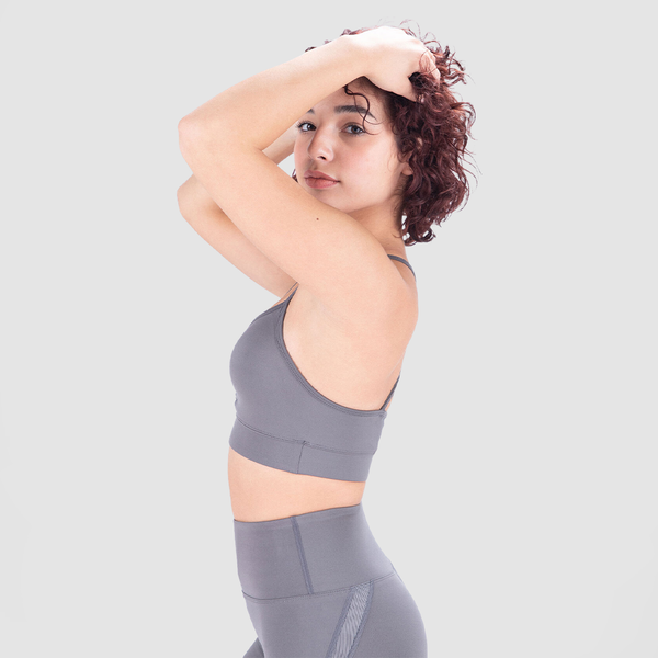 A fit woman with a friendly expression poses in a light gray athletic outfit, consisting of a sports bra and high-waisted leggings. She stands at a slight angle with her body turned to the side, smiling subtly against a plain white background