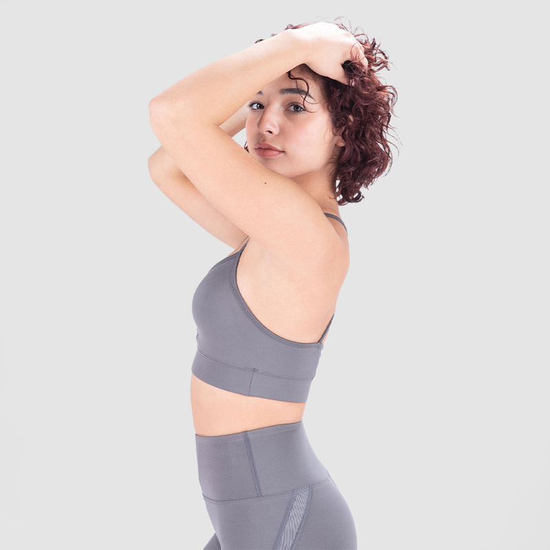 A fit woman with a friendly expression poses in a light gray athletic outfit, consisting of a sports bra and high-waisted leggings. She stands at a slight angle with her body turned to the side, smiling subtly against a plain white background