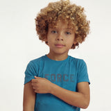 A boy posing, touching the sleeve of his t-shirt and wearing a regular fit saxony blue camo t-shirt with the word FORCE in big on it.