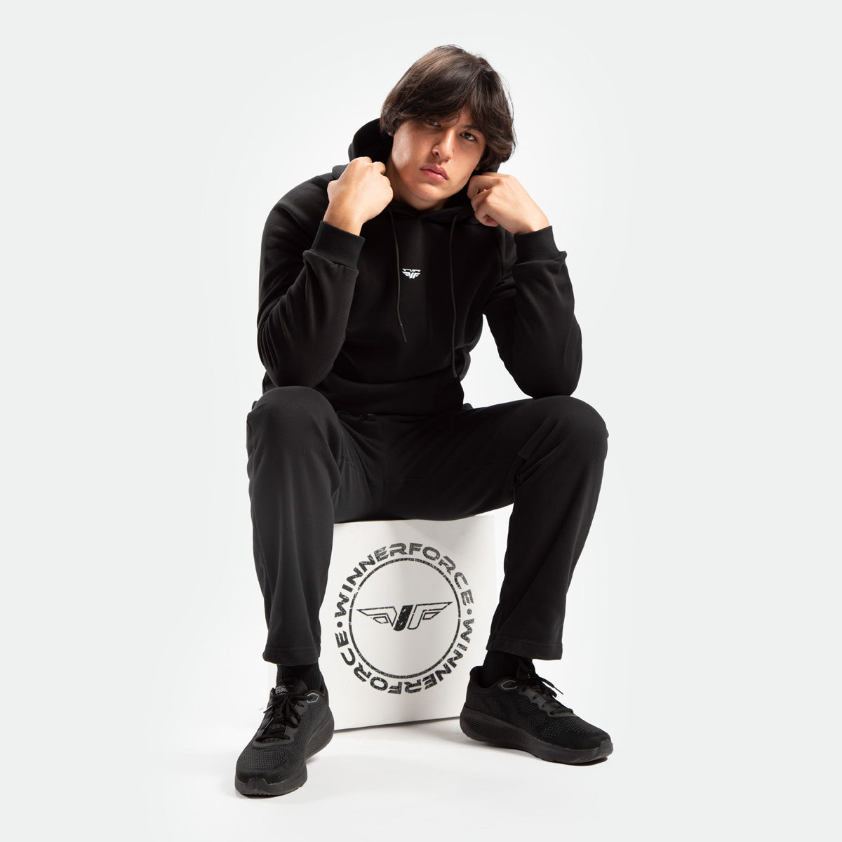 a fit man wearing matching color set black ,hoodie and pants ,plus he sits  on block white with logo winnerforce 