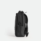 A sleek, iron grey backpack is shown standing upright on its base, viewed from the side against a plain white background. The design is minimalist with subtle zippers and padded shoulder straps visible.