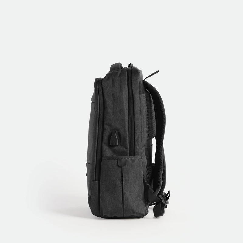 A sleek, iron grey backpack is shown standing upright on its base, viewed from the side against a plain white background. The design is minimalist with subtle zippers and padded shoulder straps visible.
