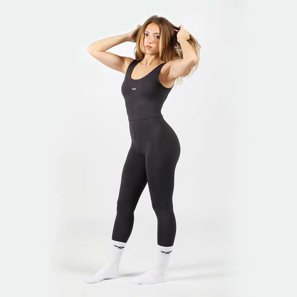a fit woman look at the camera with pose ,she wearing jump suit black with  logo socks white ,the background is light grey 