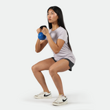 Woman performing a squat exercise while holding a blue kettlebell, wearing a light purple athletic t-shirt, black shorts, and white Nike sneakers on a light grey  background.
