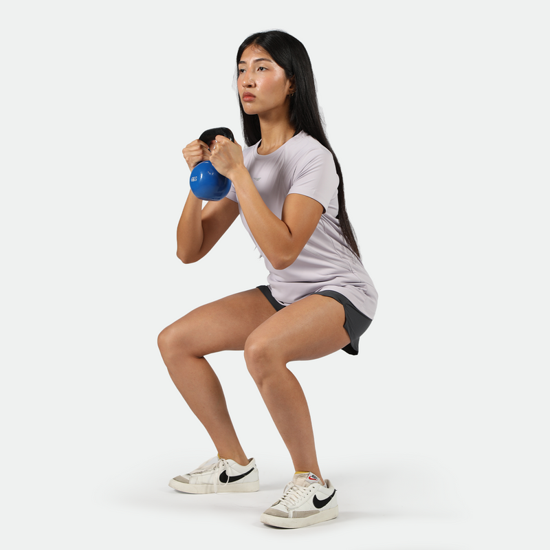 Woman performing a squat exercise while holding a blue kettlebell, wearing a light purple athletic t-shirt, black shorts, and white Nike sneakers on a light grey  background.