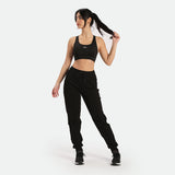 a fit woman stand at right and look at same direction ,she wearing matching black sports bras and jogger pants with shoes ,plus the background is light grey 