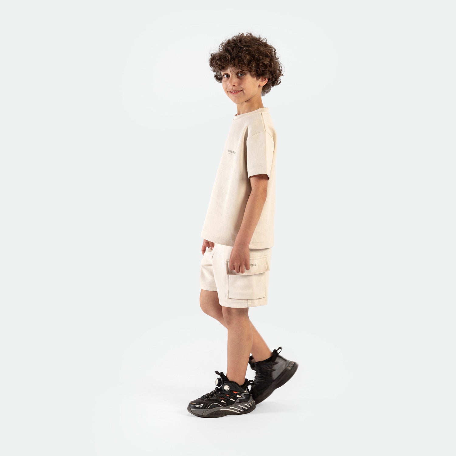 a fit boy stand at right and wearing matching set light beige ,t-shirt with short ,plus the shoes 