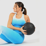 A woman with a fit physique, wearing a blue gradient sports bra and leggings, is performing a Russian twist exercise with a black medicine ball. She has her dark hair tied back in a ponytail and is looking to the side with a focused expression. The background is plain white