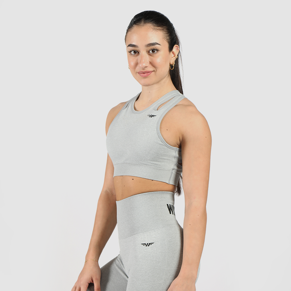 A fit woman with a friendly expression poses in a light gray athletic outfit, consisting of a sports bra and high-waisted leggings. She stands at a slight angle with her body turned to the side, smiling subtly against a plain white background