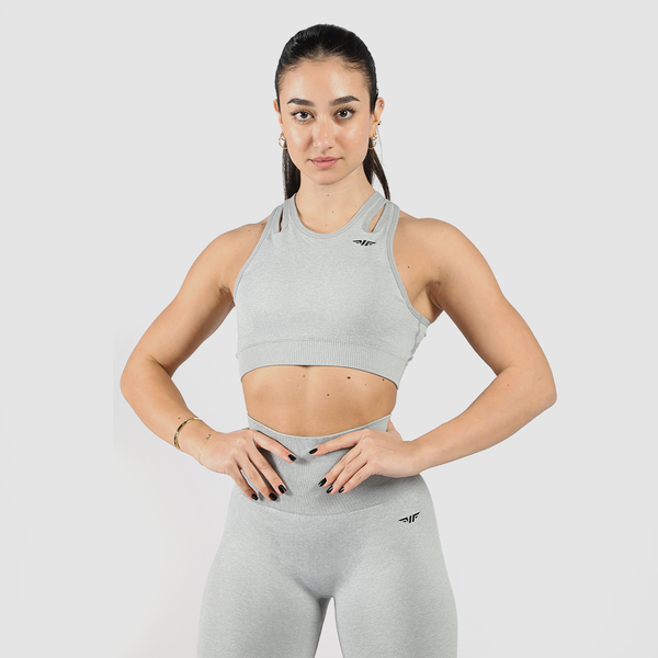 A fit woman with a confident expression poses in a light gray athletic outfit consisting of a sports bra and high-waisted leggings. She stands against a plain white background with her hands resting on her waist.