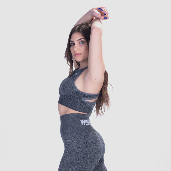 A fit woman with a friendly expression poses in a light gray athletic outfit, consisting of a sports bra and high-waisted leggings. She stands at a slight angle with her body turned to the side, smiling subtly against a plain white background