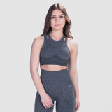A fit woman with a confident expression poses in a light gray athletic outfit consisting of a sports bra and high-waisted leggings. She stands against a plain white background with her hands resting on her waist.