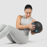 A woman in gray workout attire performs a Russian twist exercise with a black medicine ball. She is seated on the floor with knees bent, twisting her torso while holding the ball. The background is plain white