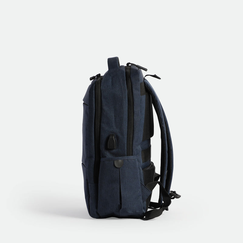 A sleek, navy blue  backpack is shown standing upright on its base, viewed from the side against a plain white background. The design is minimalist with subtle zippers and padded shoulder straps visible.