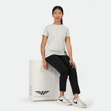 A woman in a light grey shirt and black pants sits on a white box with "Winnerforce" logo. She appears calm and confident, wearing white sneakers.