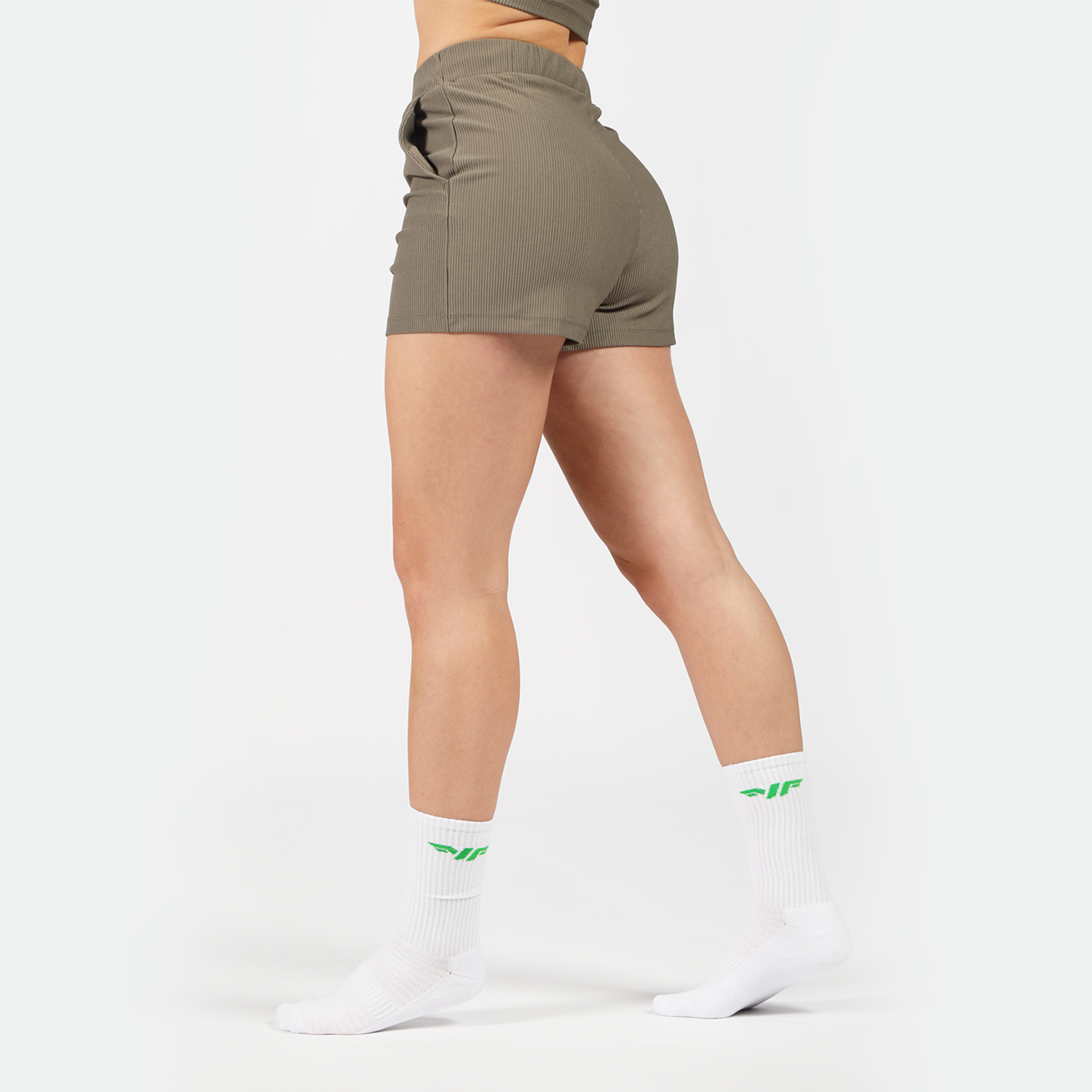 a fit woman stand at right and wearing short olive -grey with socks white ,plus the background is light grey 