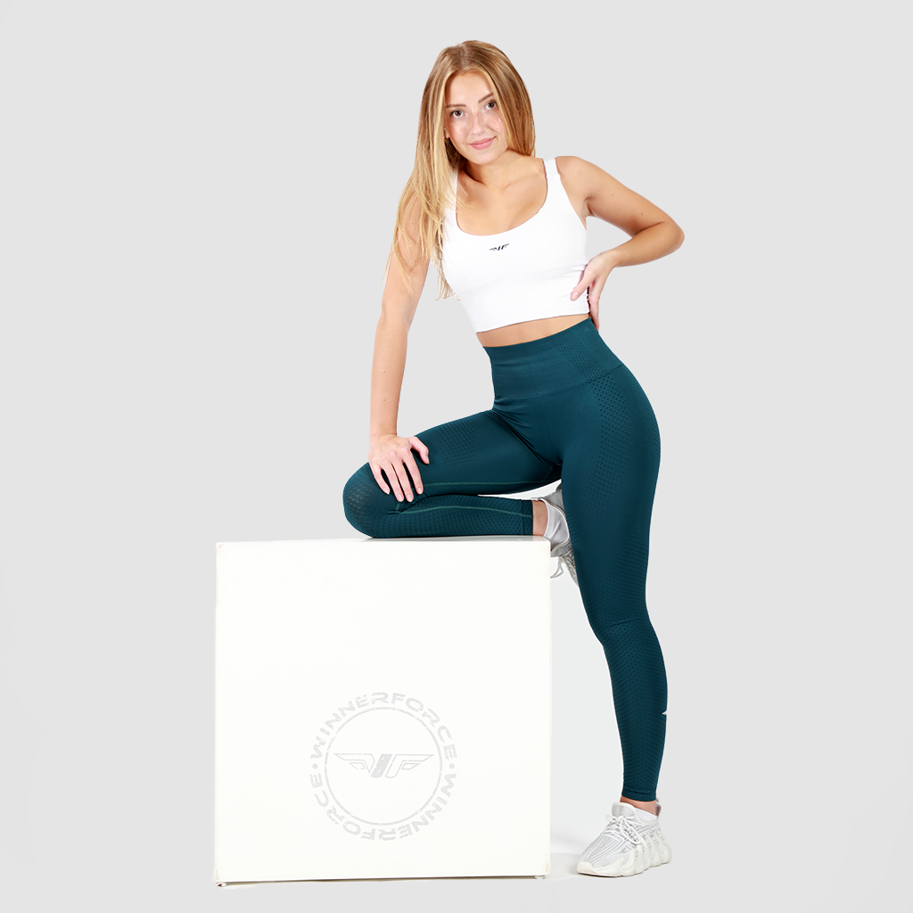 A woman with long blonde hair, wearing a white sports bra, teal high-waisted leggings, and white sneakers, poses with one hand on her hip and the other resting on a white cube. She stands confidently against a plain light gray background