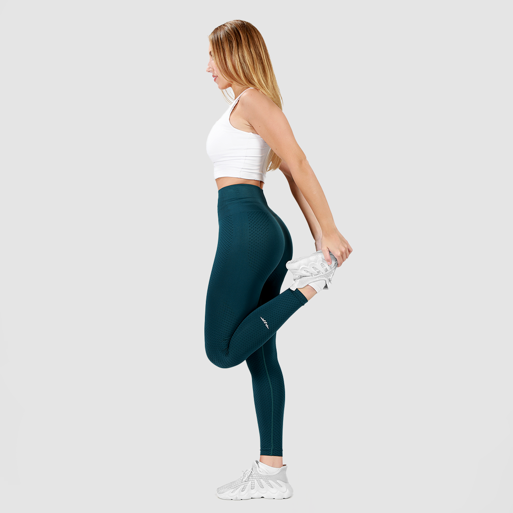 A woman with long blonde hair, wearing a white sports bra, teal high-waisted leggings, and white sneakers, performs a standing quadriceps stretch. She balances on one leg while pulling her other foot towards her glutes, looking straight ahead against a plain light gray background