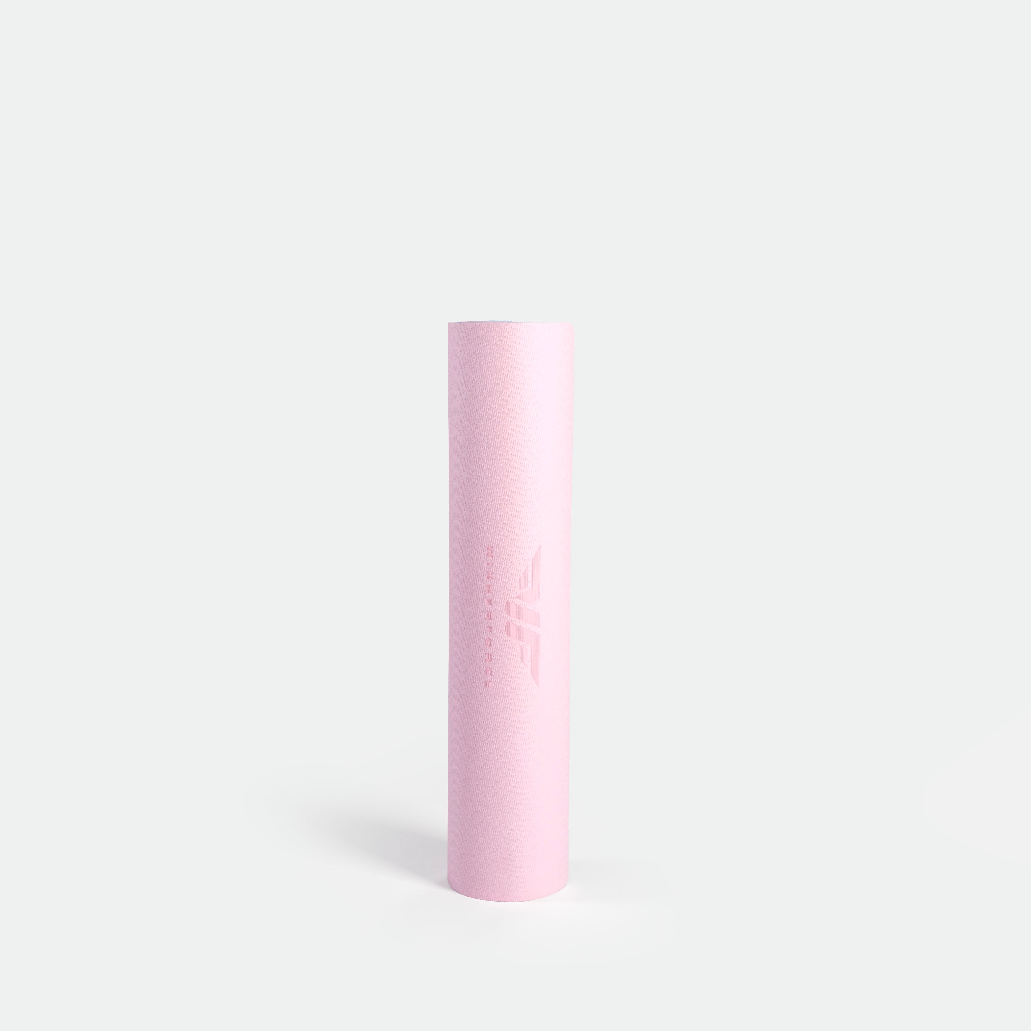 A tall, cylindrical, pastel pink yoga mat against a plain white background. The minimal design conveys simplicity and elegance, with a subtle logo.
