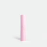 A tall, cylindrical, pastel pink yoga mat against a plain white background. The minimal design conveys simplicity and elegance, with a subtle logo.